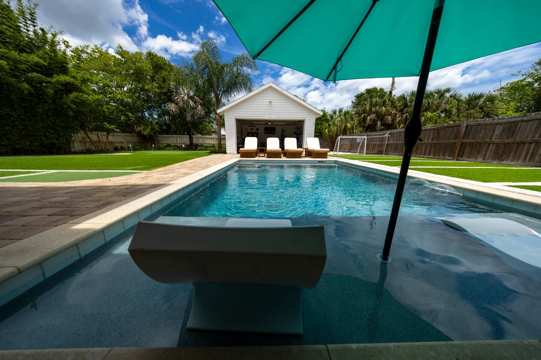 Heated pool with in-water loungers and a teal umbrella, backed by lounge chairs, sports turf, and a garage game room at Family Tides