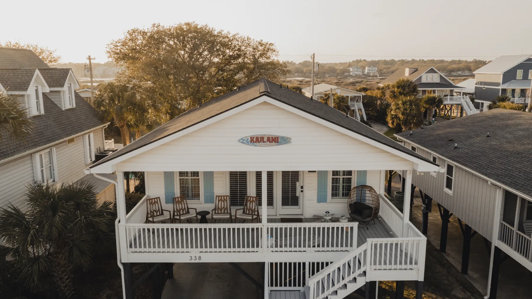Aerial view of Kailani House, a raised white beach cottage with a wide front porch, at golden hour in Murrells Inlet, South Carolina